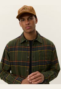 Groene geruite flanellen shirt met gele en rode accenten, voorzien van een kraag en een knoopsluiting. G gedragen met een bruine corduroy pet.