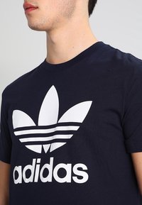 Námořnickě modré bavlněné tričko s bílou grafikou tří tvarů listů a slovem "adidas" v tučném, čistém písmu níže.
