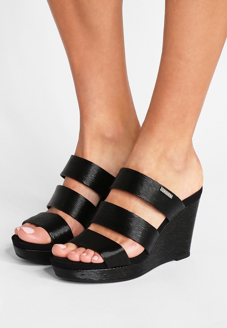 Des pieds portant des sandales compensées noires avec trois larges sangles texturées sur les orteils et le dessus du pied, sur un fond blanc.