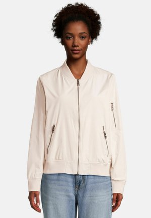 Vrouw draagt een lichtbeige bomberjack met ritssluitingen en ritssluitingen op de zakken, gecombineerd met lichtblauwe spijkerbroek, staand voor een effen achtergrond.