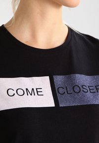 Žena v černém tričku s třpytivými bloky a nápisy "PŘIJDI" a "BLÍŽ" tištěnými přes hrudník.