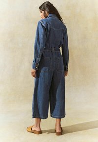 Jeansoverall med midjebälte, långa ärmar, vida ben och två bakfickor. Färgen är mörkblå med en klassisk denimtextur.