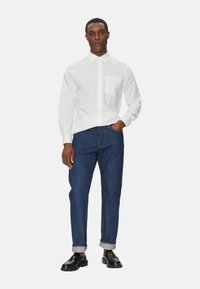Camicia bianca con maniche lunghe e taschino sul petto, abbinata a jeans diritto blu scuro e scarpe nere. Design semplice e pulito.