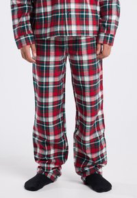 Child wearing red, green, and white plaid pajama pants and matching long-sleeve top, standing with black socks against a plain background.