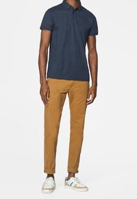 Polo shirt bleu marine avec un motif texturé, pantalon slim-fit kaki, et baskets blanches avec des accents bleus, se tenant devant un fond blanc.