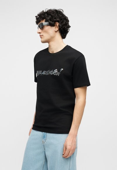 Νέος άνδρας με σγουρά μαλλιά, φορώντας μαύρο μπλουζάκι με σχέδιο, ανοιχτό μπλε τζιν και ορθογώνια μαύρα γυαλιά ηλίου, στραμμένος προς τα αριστερά σε απλό φόντο.