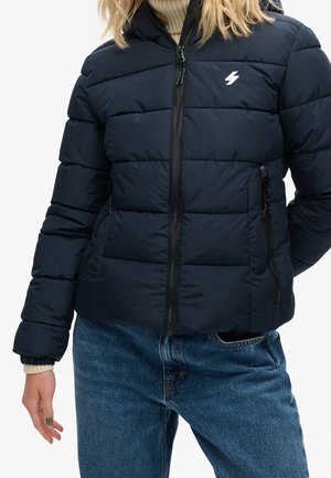 Femme portant une veste matelassée bleu marine avec des fermetures éclair à l'avant et sur les côtés, un petit logo éclair blanc, et un jean bleu.