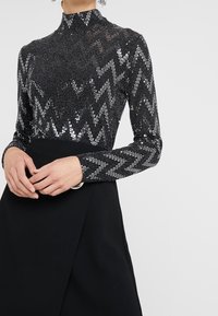 Svart långärmad topp med silverpaljetter i zigzag-mönster, hög krage. Kombinationen med en svart kjol med omslagsdesign och cirkulär bältesdetalj.