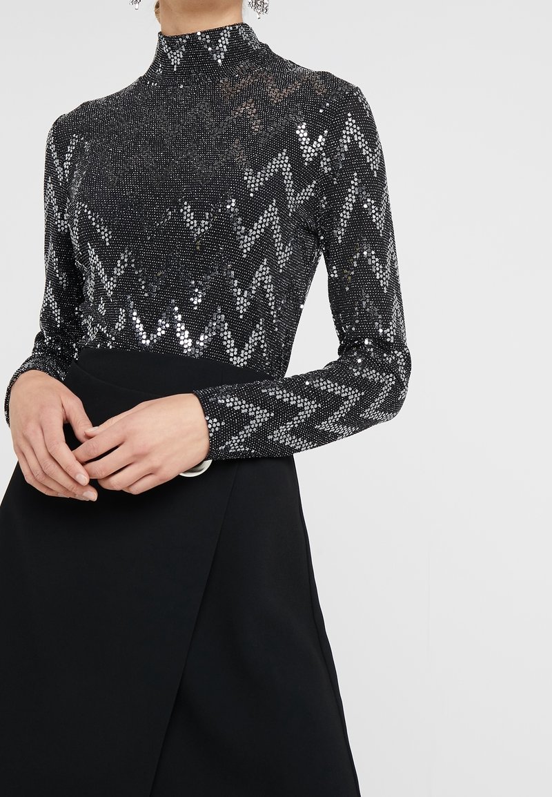 Svart långärmad topp med silverpaljetter i zigzag-mönster, hög krage. Kombinationen med en svart kjol med omslagsdesign och cirkulär bältesdetalj.