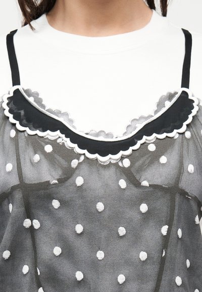 Κοντινή λήψη μαύρου διαφανούς φορέματος με πουά και λευκή κυματιστή διακόσμηση πάνω από ένα απλό λευκό τοπ με στρογγυλή λαιμόκοψη.