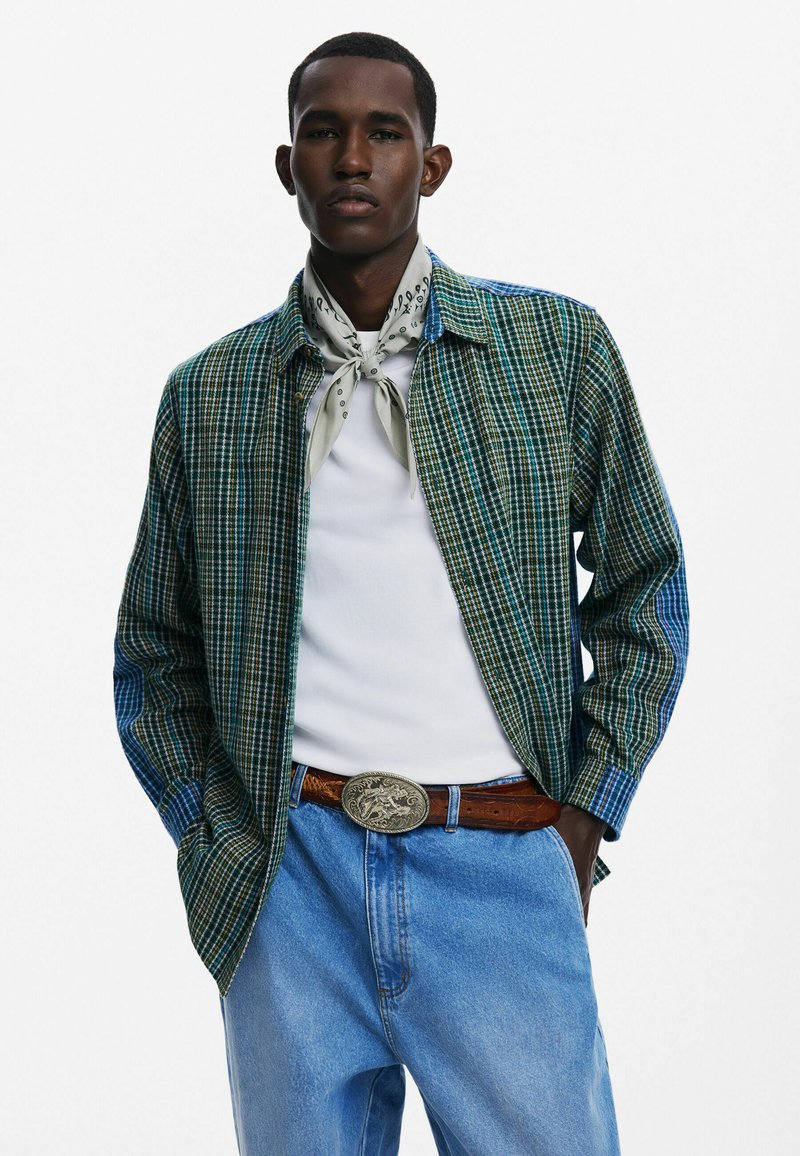 Koszula w kolorową kratę w odcieniach zieleni i niebieskiego, dżinsowe spodnie, warstwowe z białą koszulą i lekkim szalikiem, z metalową klamrą paska w kolorze srebrnym.