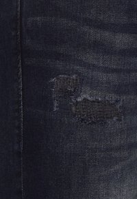 Tessuto di jeans blu scuro con due toppe scucite ai bordi. La texture è liscia con una leggera lucentezza.