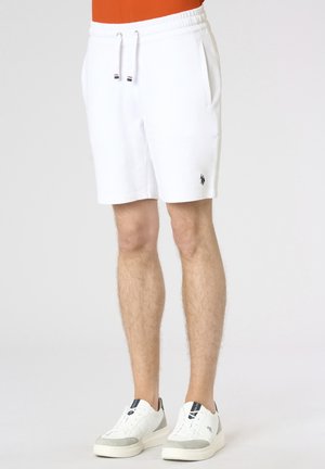 Uomo che indossa pantaloncini bianchi con coulisse e tasche laterali e sneakers bianche con dettagli grigi su sfondo bianco.