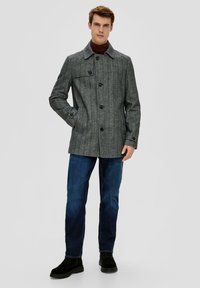 Grau strukturierter Mantel mit spitzem Kragen und vier vorderen Knöpfen, kombiniert mit dunkelblauen Jeans und schwarzen Wildlederschuhen.