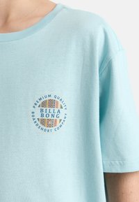 T-shirt in cotone azzurro chiaro con scollatura rotonda e maniche corte. Presenta un logo circolare "BILLA BONG" con motivi colorati.