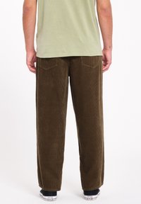 Pantalons en velours côtelé marron avec des jambes larges, des poches arrière et une texture à côtes verticales. Portés avec un t-shirt vert et des baskets noires.