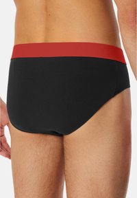 Sorte svømmedragter med en rød elastisk talje, der har en glat tekstur og et tætsiddende design, som viser minimale syningsdetaljer.