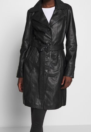 Trench-coat en cuir noir avec ceinture et boutons, porté sur un haut blanc et un pantalon noir, longueur mi-cuisse, poches avant et côté visibles.