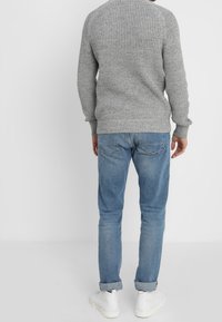Pull en tricot gris texturé avec un col rond, associé à un jean en denim bleu clair et des baskets blanches. Détails visibles au dos et ajustement.