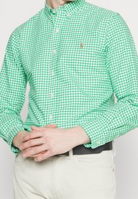 Zelená a bílá kostkovaná košile na knoflíky s klasickým límečkem, s malým logem na levé straně hrudi, v kombinaci se světlými kalhotami.