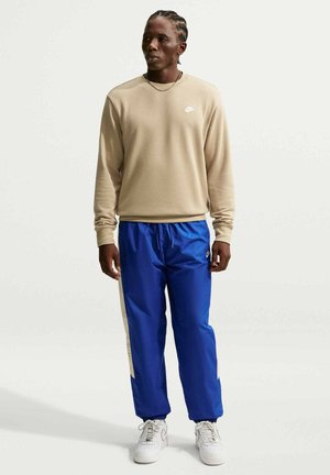 Man met gevlochten haar draagt een beige Nike-trui, blauwe Nike trainingsbroek en witte sneakers, staand op een effen witte achtergrond.