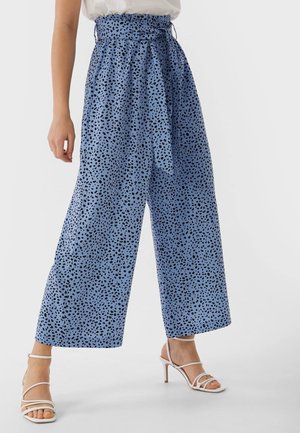 Femme portant un pantalon large bleu taille haute avec des taches noires, ceinture nouée, sandales blanches à talons avec des brides, et un haut blanc rentré.