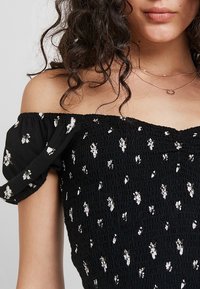 Top negro sin hombros hecho de tela fruncida que presenta pequeños patrones florales blancos y mangas abullonadas. Textura suave con un ligero brillo.