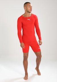Červená sportovní rash guard s dlouhými rukávy, na tělo přiléhající design a bílý logo na hrudi. V páru s odpovídajícími červenými kompresními šortkami.