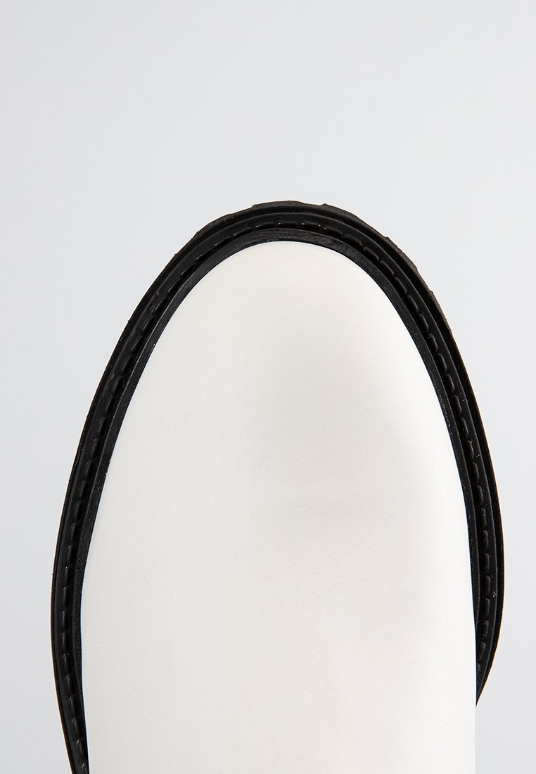 Stivaletto in pelle bianca con profilo in gomma nera, caratterizzato da una superficie liscia e una forma arrotondata nella parte anteriore.