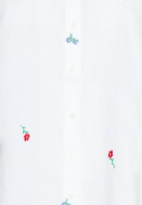 Chemise blanche ornée d'une broderie florale colorée en bleu et rouge. Elle présente un modèle à boutons sur le devant avec des boutons blancs et un tissu texturé.