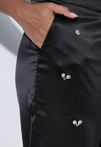 Černá saténová sukně s boční kapsou, zdobená třemi průhlednými kamennými ozdobami různých velikostí připevněnými na látce.