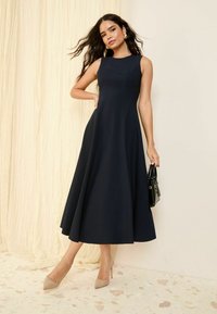 Navy A-line dress with a fitted bodice and flared skirt, made of soft fabric. Model wears nude pointed heels and holds a black handbag.