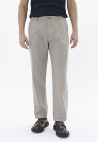 Pantalones beige ajustados con corte recto, pliegues y cierre de botón. La tela tiene una superficie texturizada. Se combinan con zapatos marrón oscuro.