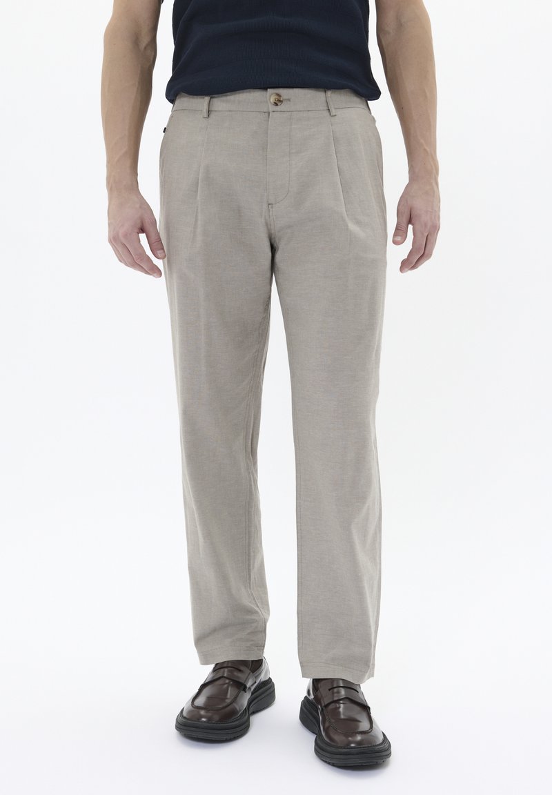 Pantalones beige ajustados con corte recto, pliegues y cierre de botón. La tela tiene una superficie texturizada. Se combinan con zapatos marrón oscuro.
