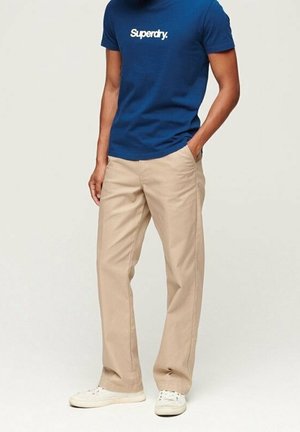 Un pantalon chino beige avec une coupe droite, associé à un t-shirt bleu marine avec un logo blanc. Porté avec des baskets blanches.