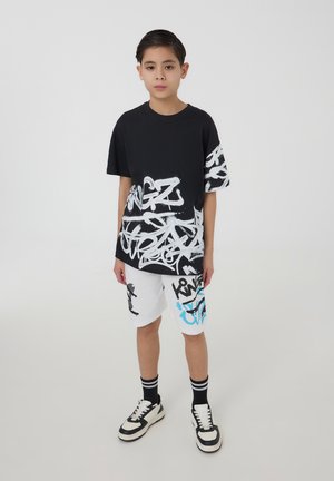 T-shirt nera con stampa graffitita bianca abbinata a pantaloni corti bianchi con accenti graffititi neri e blu; sneakers in bianco e nero.
