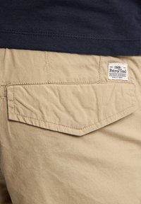 Beige broek met een achterzak die een diagonale flap en een merklabel toont, met een textuurstof en zichtbare stikdetails.