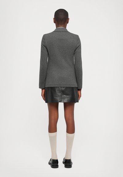 Grijze, gestructureerde blazer met een getailleerde pasvorm, gedragen over een zwarte leren rok. Het model heeft kniehoge beige sokken en zwarte schoenen aan.