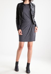 Schwarze Lederbikerjacke, die über einem grauen kurzärmeligen Kleid mit verdrehtem Vorderteil getragen wird, ergänzt durch schwarze Knöchelstiefel.