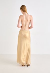 Goldfarbene Satin-Halterneckkleid mit offenem Rücken, gerafftem Detail an der Taille und bodenlanger fließender Form, kombiniert mit schwarzen High Heels.