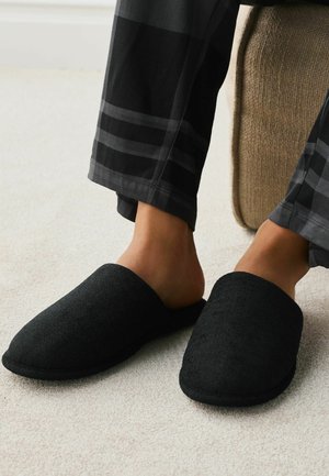 Person trägt schwarze weiche Stoff-Pantoffeln zum Hineinschlüpfen und schwarz karierte Pyjamahosen, sitzt auf einem beigen Stuhl auf hellem Teppich.