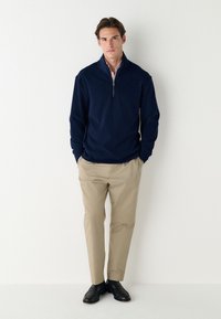 Námořnický svetřík na zip, béžové kalhoty a černé boty. Svetr má vysoký límec a volný střih. Hladká textura a klasický design.