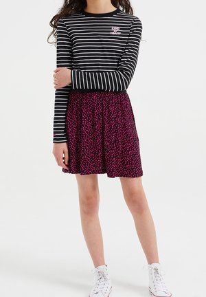 Pige iført sort-hvid stribet langærmet trøje med teksten "KIND HEART", lilla nederdel med lyserøde pletter og hvide sneakers.