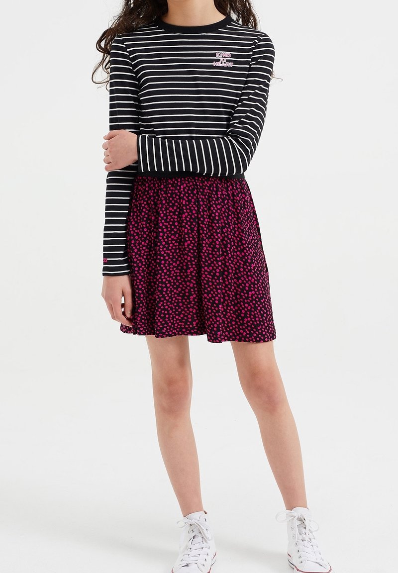 Dziewczyna ubrana w czarno-biały pasiasty, długi rękaw z napisem "KIND HEART", fioletową spódnicę w różowe kropki oraz białe tenisówki.