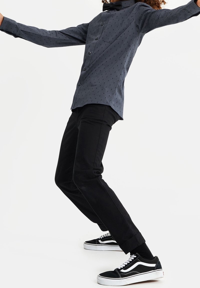 Dlouhý rukáv tmavě šedé košile na knoflíky s drobným vzorem, kombinované se slim-fit černými kalhotami a černými teniskami Vans.