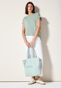 Haut à manches courtes à rayures vertes et blanches, pantalon blanc, tenant un sac fourre-tout vert menthe avec "CECIL" imprimé. Chaussures beiges avec des accents blancs.