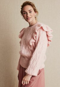 Różowy sweter dzianinowy z falbaniastymi rękawami, z wzorem w warkocze. Miękka tekstura, dopasowany krój, w komplecie z różową spódnicą.