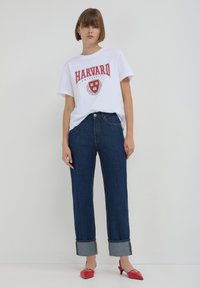 T-shirt en coton blanc avec le texte "HARVARD" rouge, associé à un jean en denim bleu ample retroussé à l'ourlet, et des chaussures à talons rouges.