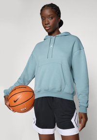 Ljusblå huvtröja med känguruficka, kombinerad med svarta shorts med vita accenter. Modellen håller en orange basketboll.
