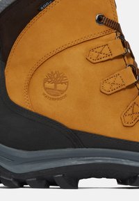 Botte de randonnée imperméable avec une tige en cuir tan, semelle en caoutchouc gris foncé et œillets en métal. Présente un logo d'arbre embossé sur le côté.